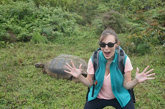 Caiti wearing a pink shirt, green vest and sunglasses, looking shocked and excited with a giant tortoise in the background